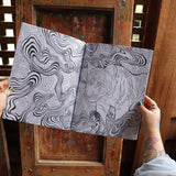 Someone holding the Psychedelic Cats Coloring Book open in front of a wooden background, showing off the pages.