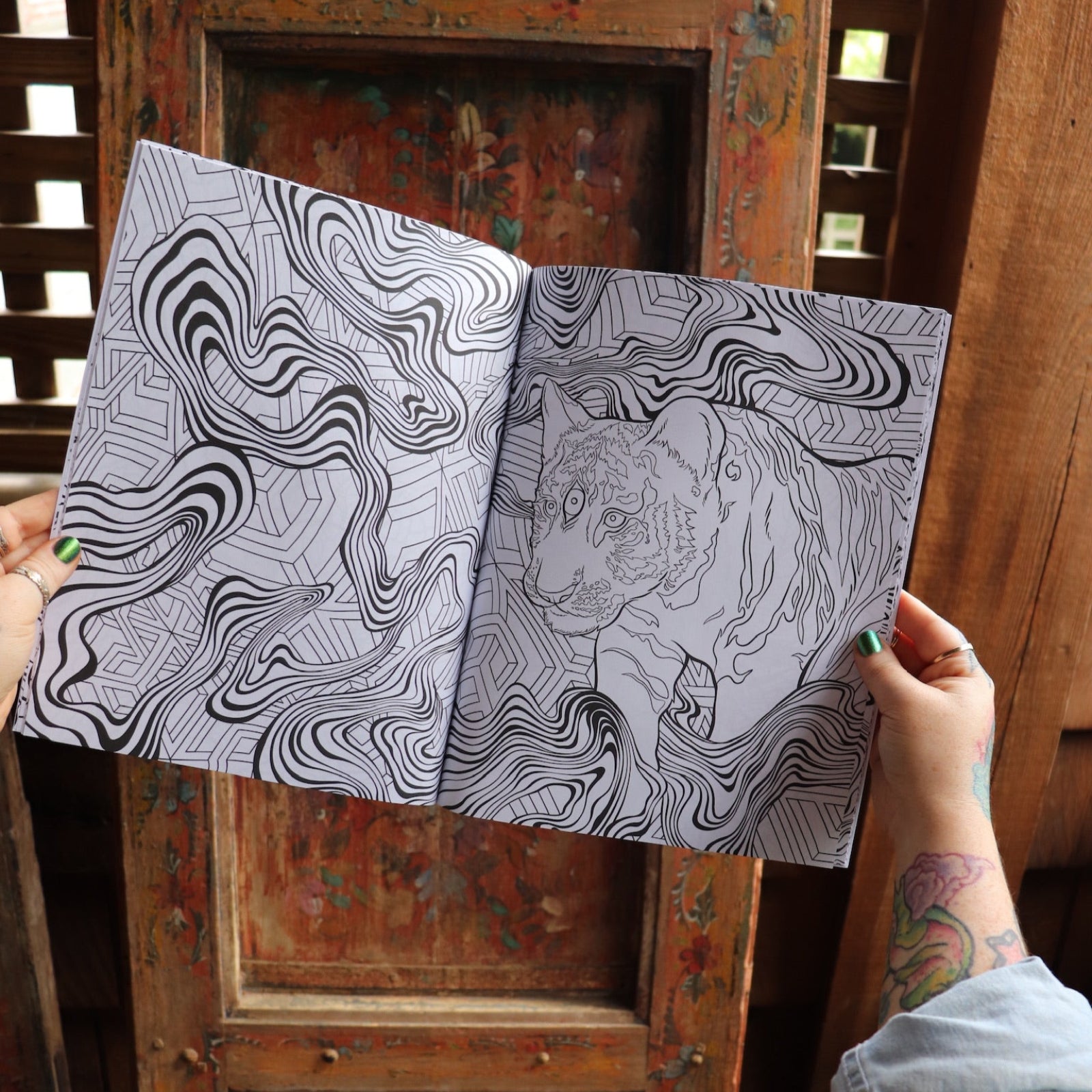 Someone holding the Psychedelic Cats Coloring Book open in front of a wooden background, showing off the pages.