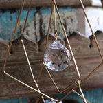 An up-close image of the Prisma Hanging #27 Brass Suncatcher in front of a wooden background, showing off a crystal.