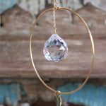 An up-close image of someone holding the Prisma Hanging #22 Brass Round Suncatcher in front of a wooden background, showing off the main crystal.