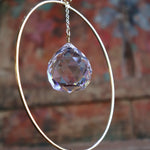 An up-close image of someone holding the Prisma Hanging #22 Brass Round Suncatcher in front of a wooden background, showing off the main crystal.