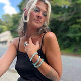 Woman with blonde hair and tattoos wearing a black tank top and turquoise bracelets outdoors.