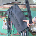 Black leather clutch handbag with fringe details held by a person against a rustic background.