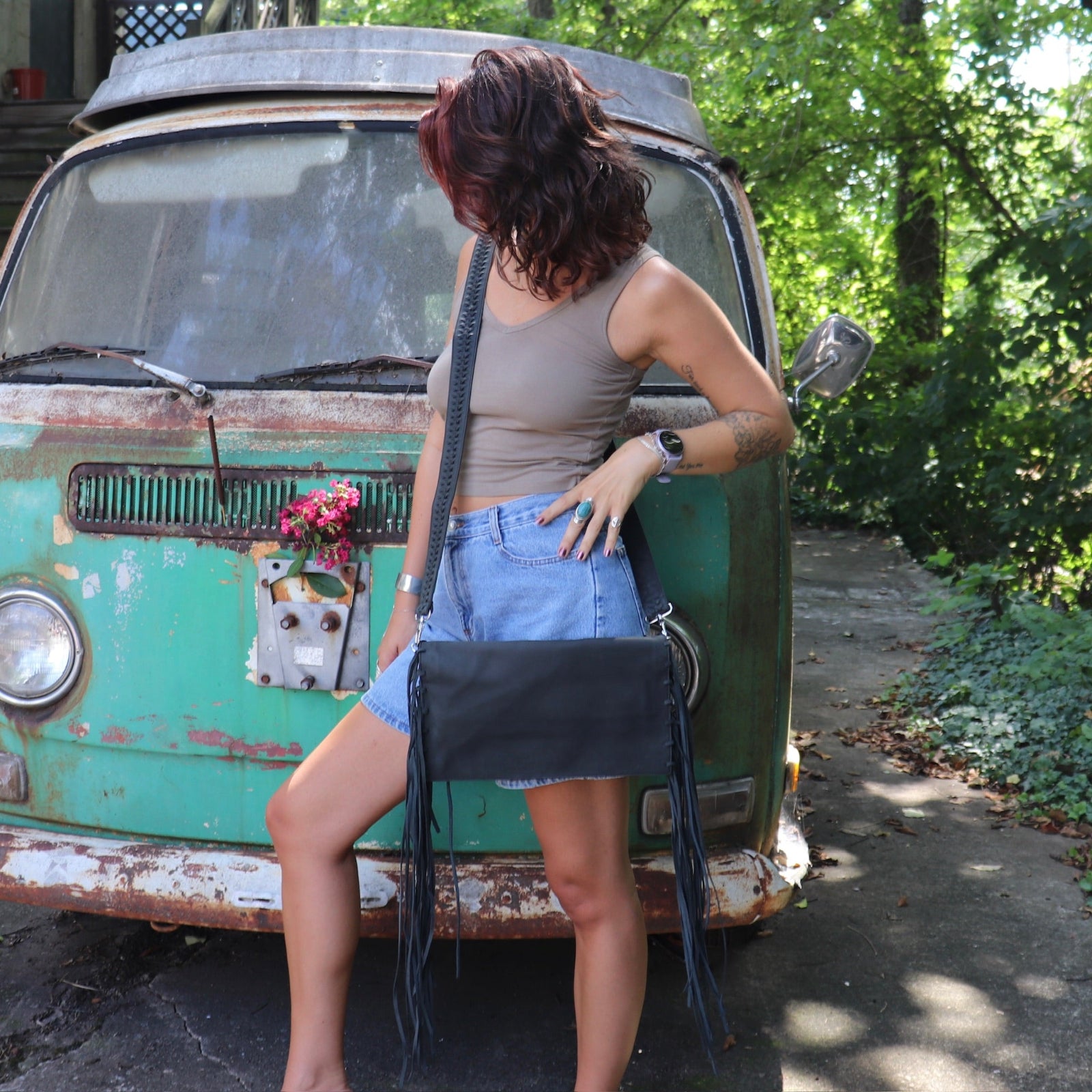 Woman leaning against a vintage green van with a black leather clutch handbag, surrounded by greenery.