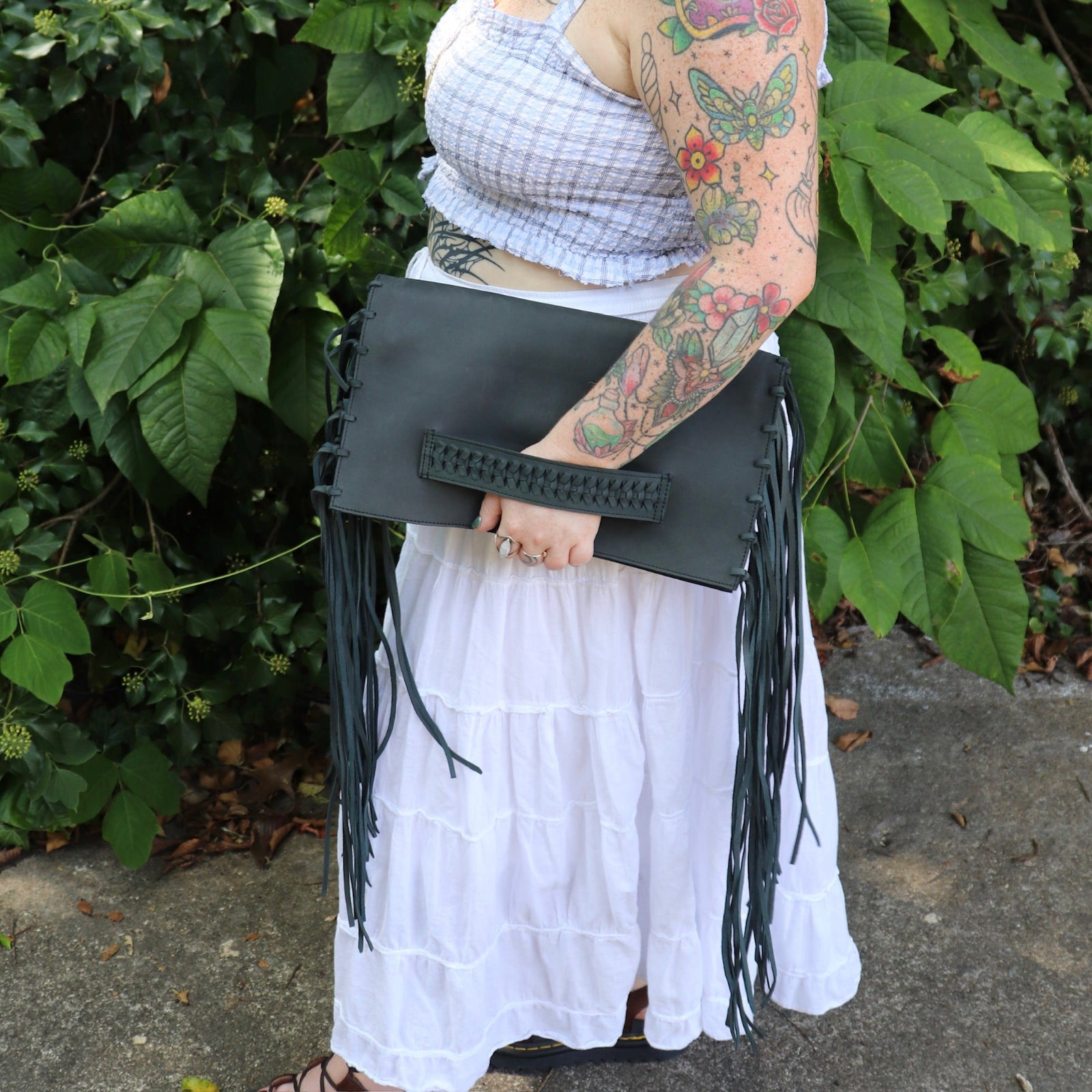 Person holding a black fringe handbag with a white skirt and tattooed arm against a green leafy background