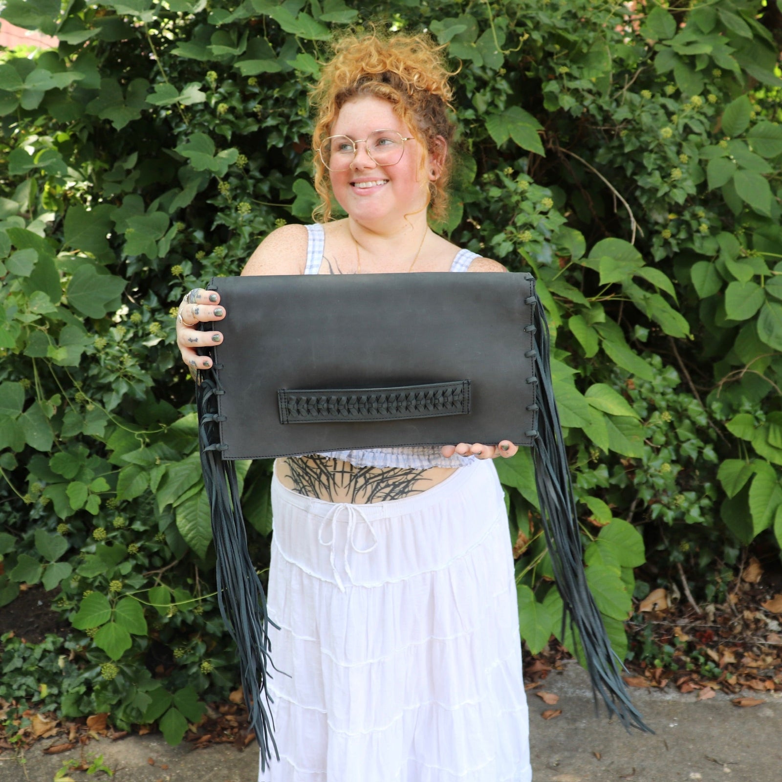 Person holding a black leather clutch with fringe details against a green leafy background