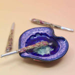 Holographic package of Manifest Magic Rollies with visible herbs inside, surrounded by individual rollies on a soft blush background