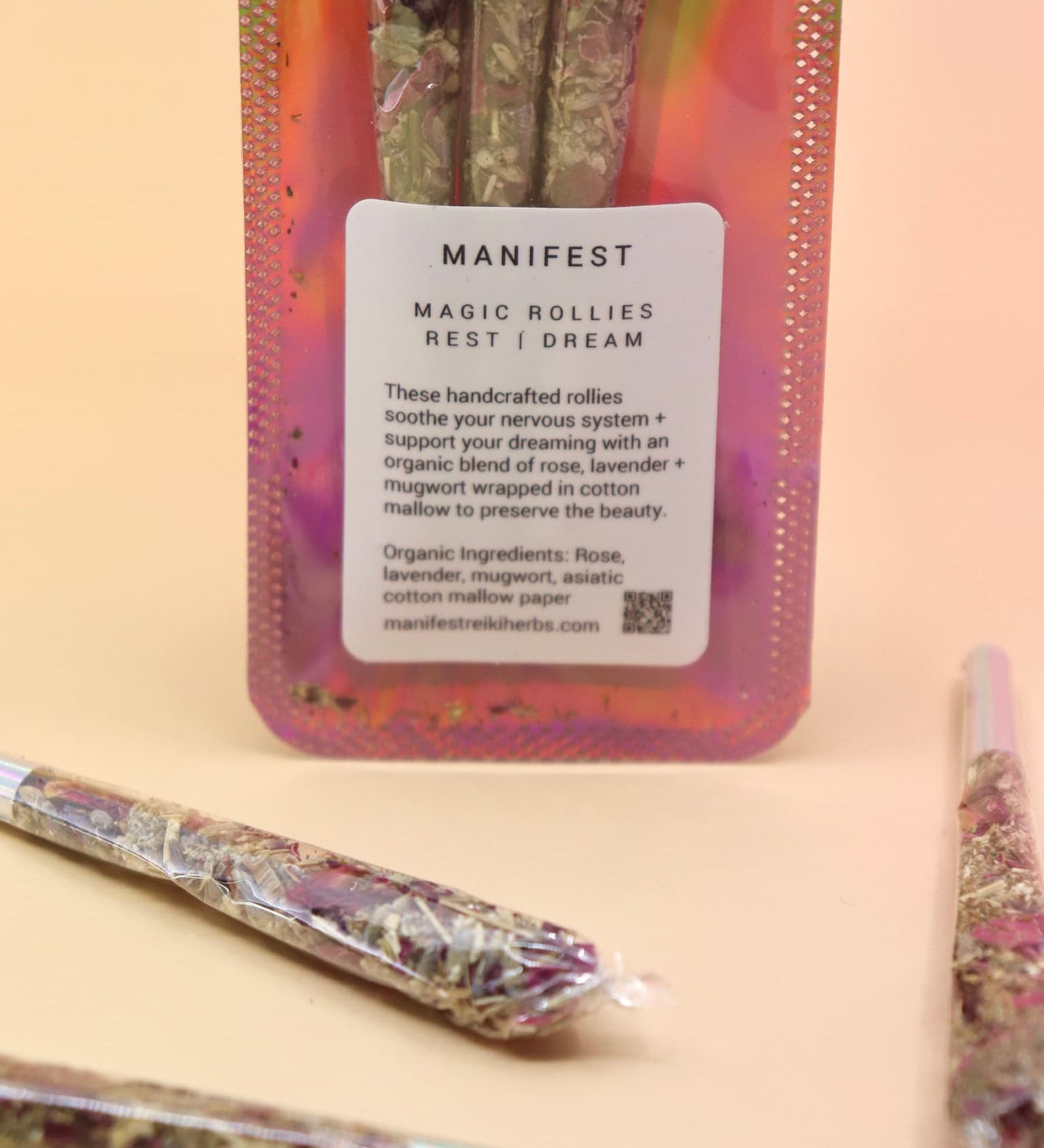 Holographic package of Manifest Magic Rollies with visible herbs inside, surrounded by individual rollies on a soft blush background