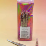 Holographic package of Manifest Magic Rollies with visible herbs inside, surrounded by individual rollies on a soft blush background