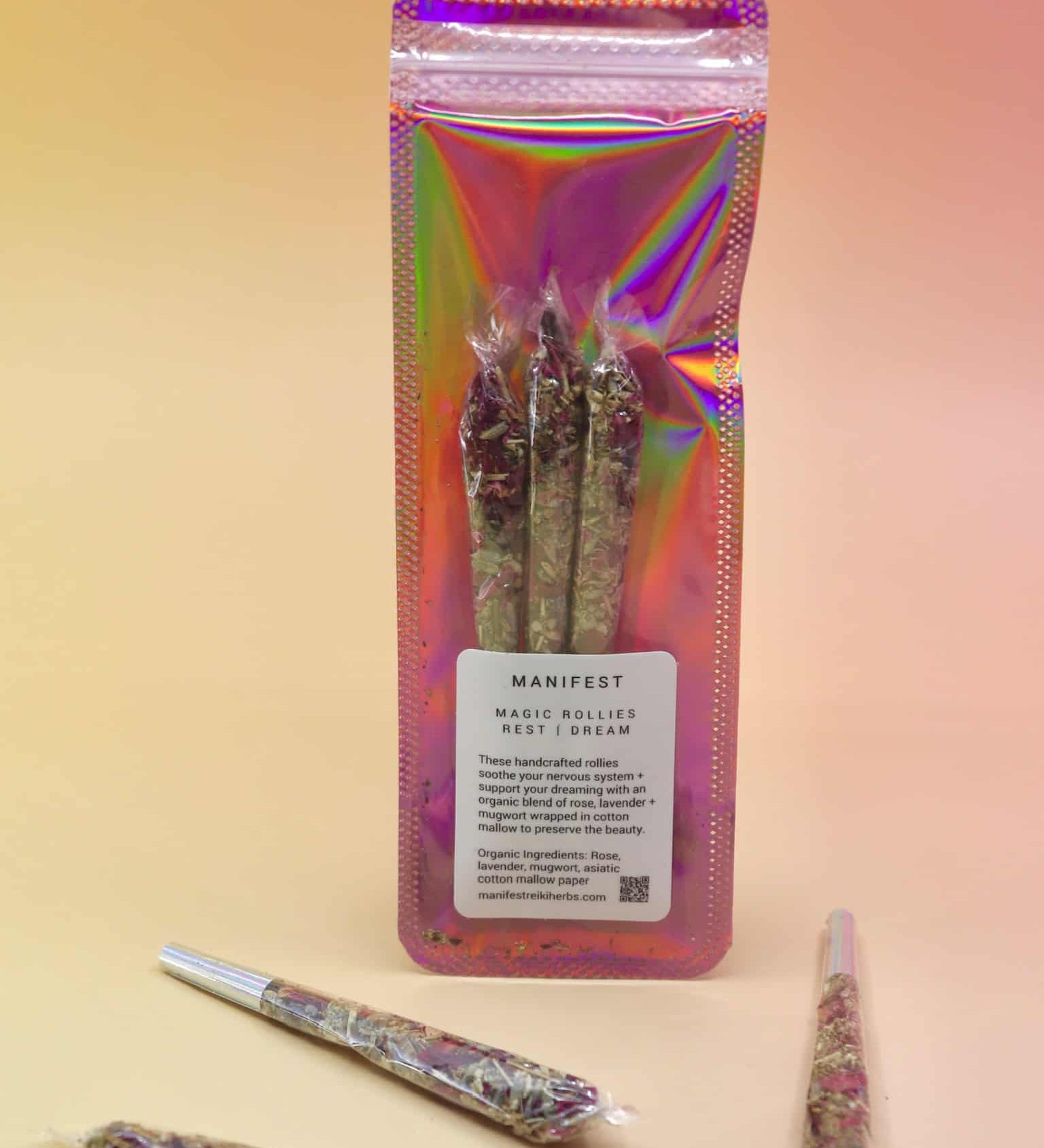 Holographic package of Manifest Magic Rollies with visible herbs inside, surrounded by individual rollies on a soft blush background