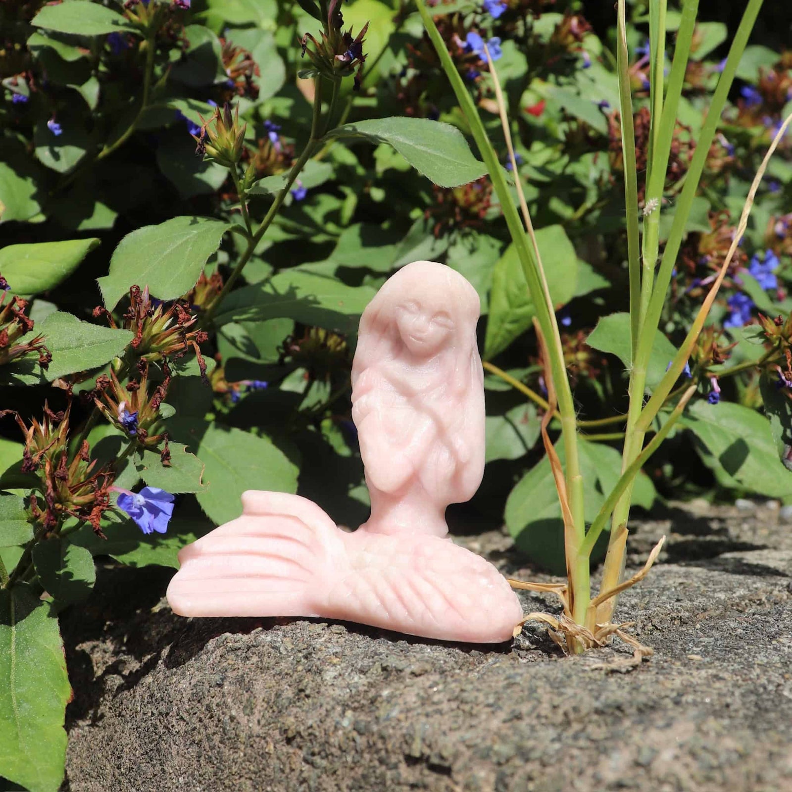 Pink opal crystal stone mermaid sculpture of a reclining figure on a rock with greenery and flowers in the background