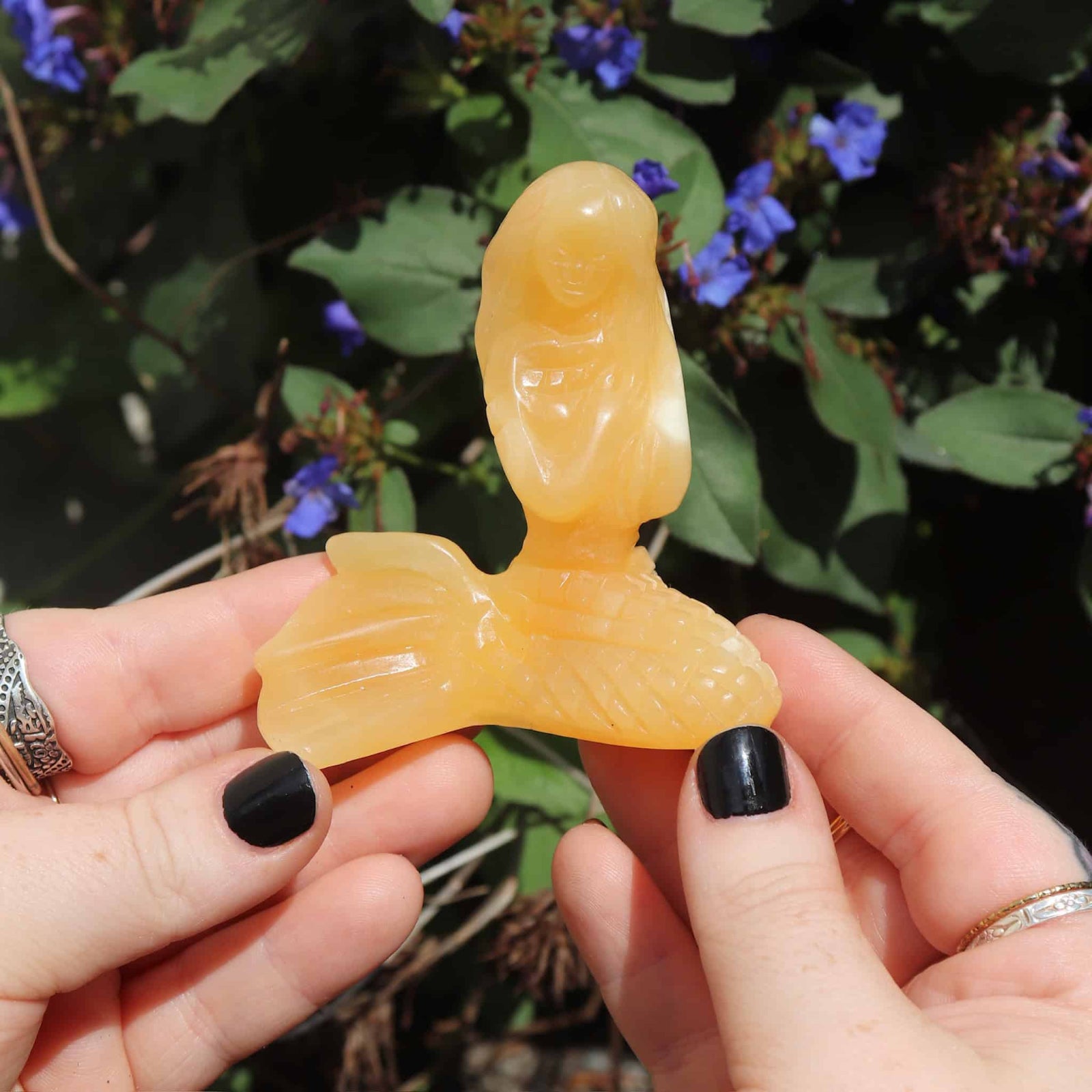 Hand holding an orange stone sculpture of a mermaid against a natural background with green leaves and blue flowers.