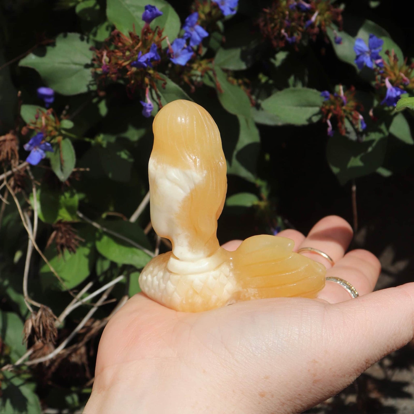 Hand holding an orange-colored mermaid crystal against a natural background with green leaves and purple flowers.