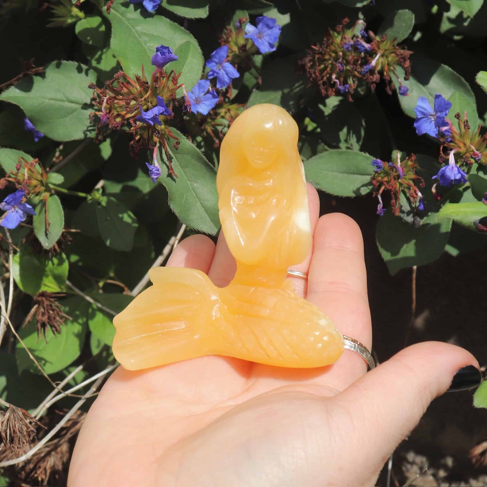 Hand holding an orange mermaid crystal sculpture against a natural background with green leaves and purple flowers.
