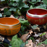 Two singing bowls, one orange and one red, with engraved text, on a natural background of leaves and earth.