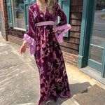 A woman wearing the 1960s Purple Floral Burnout Velvet Maxi Dress in front of a store front background.