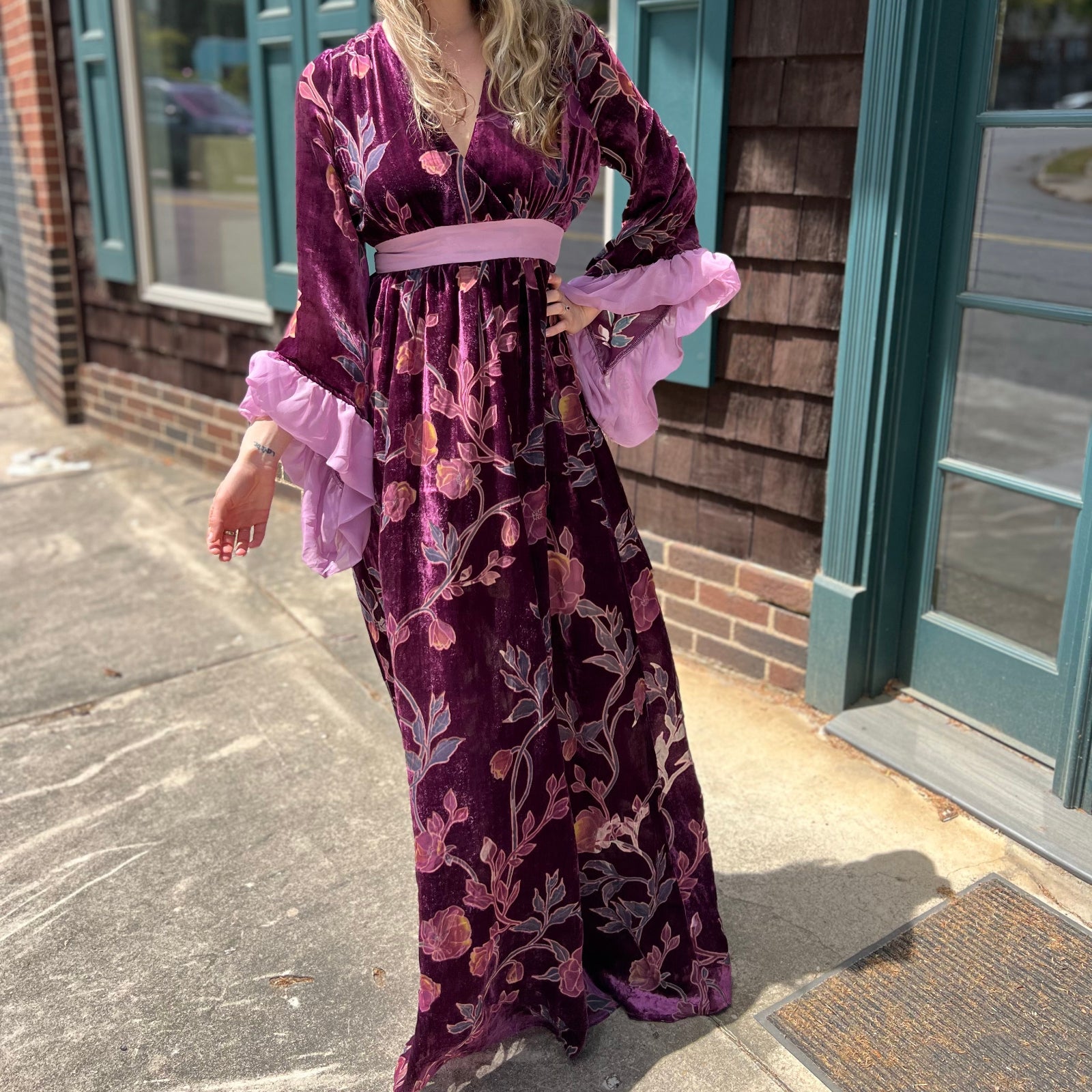 A woman wearing the 1960s Purple Floral Burnout Velvet Maxi Dress in front of a store front background.