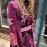 A woman wearing the 1960s Purple Floral Burnout Velvet Maxi Dress in front of a store front background, showing off the side.