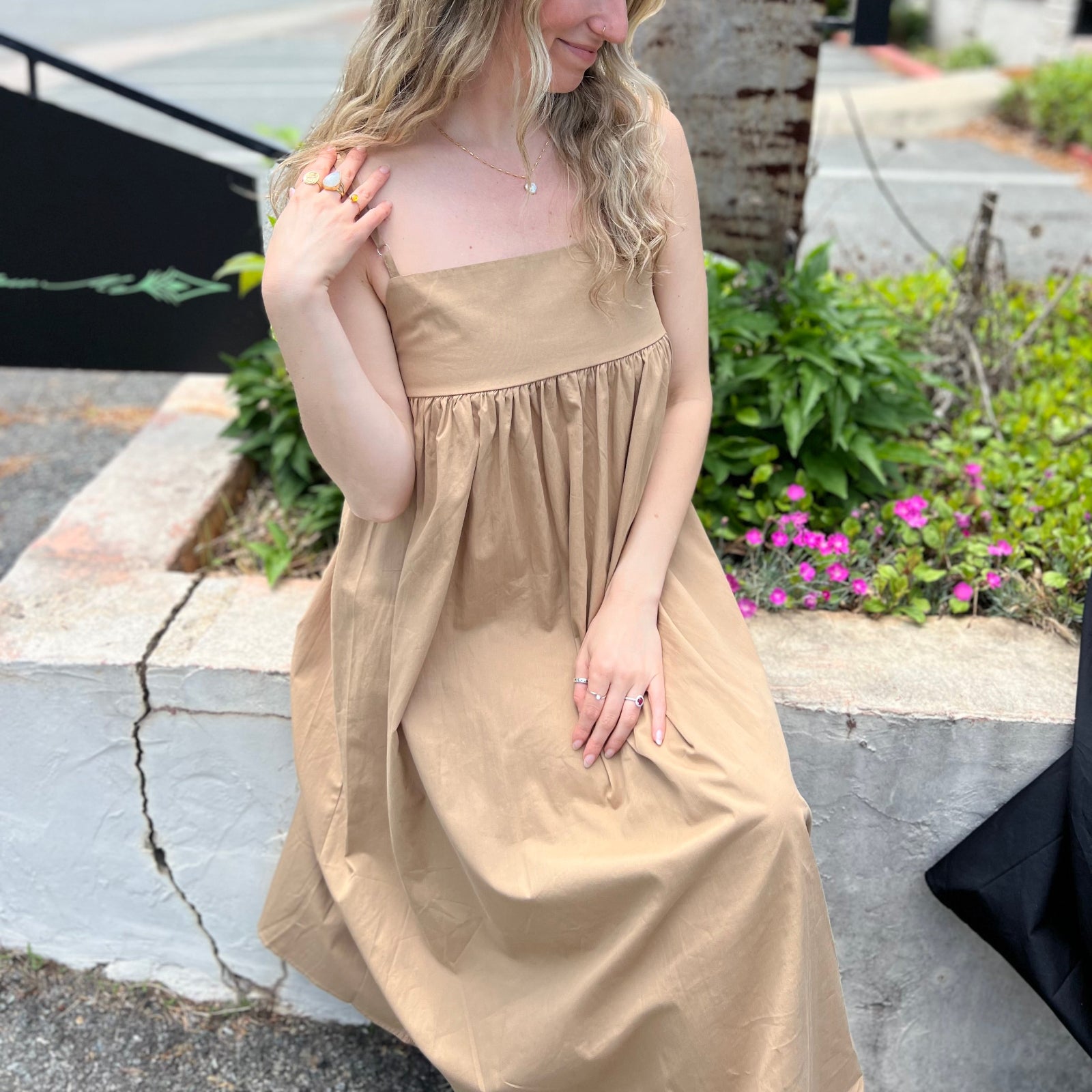 A woman wearing the khaki Solid Color Pleats Suspender Acetate Fiber Free Size Dress while sitting on a white ledge.