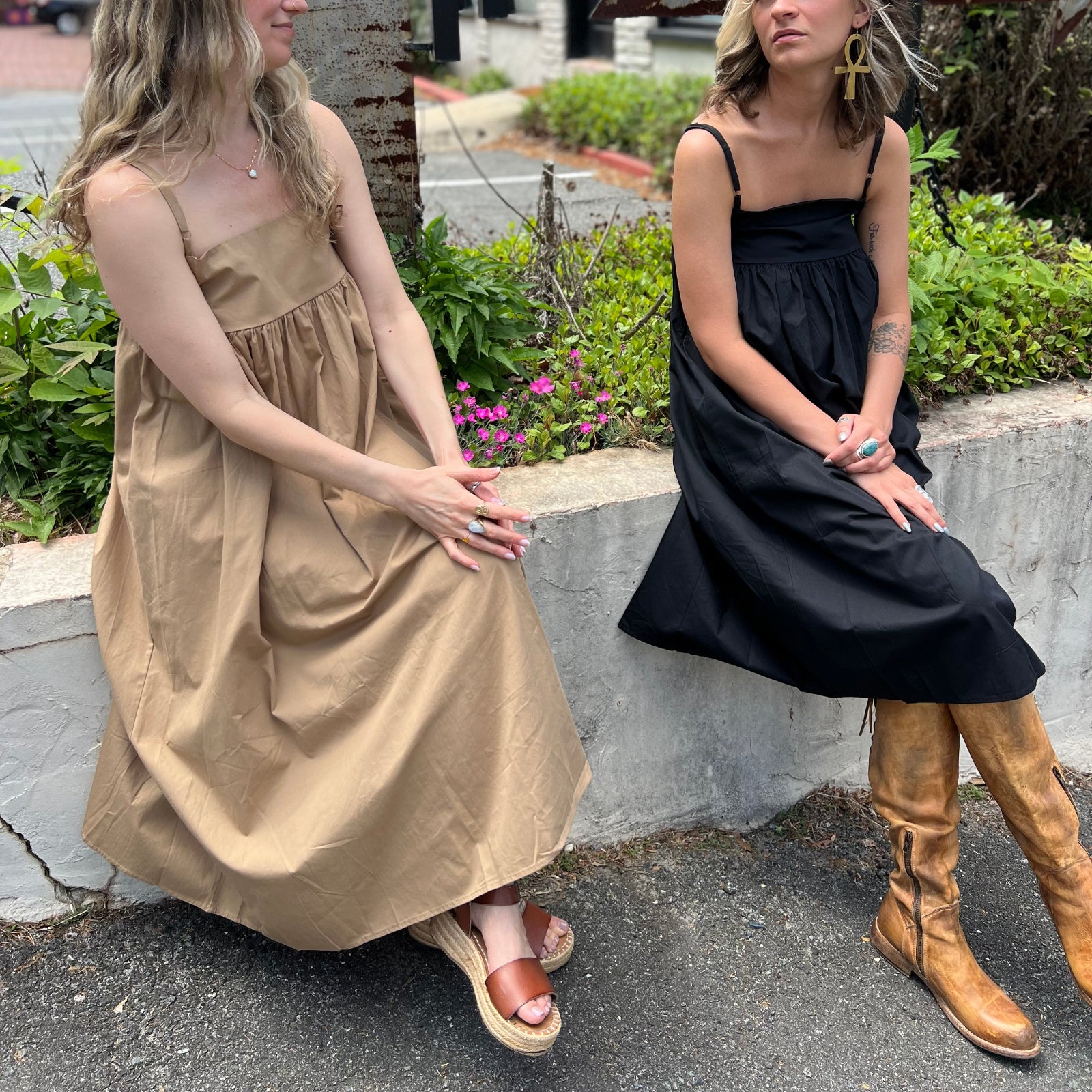 Two women wearing the Solid Color Pleats Suspender Acetate Fiber Free Size Dresses in black and khaki while sitting on a white ledge in front of greenery.