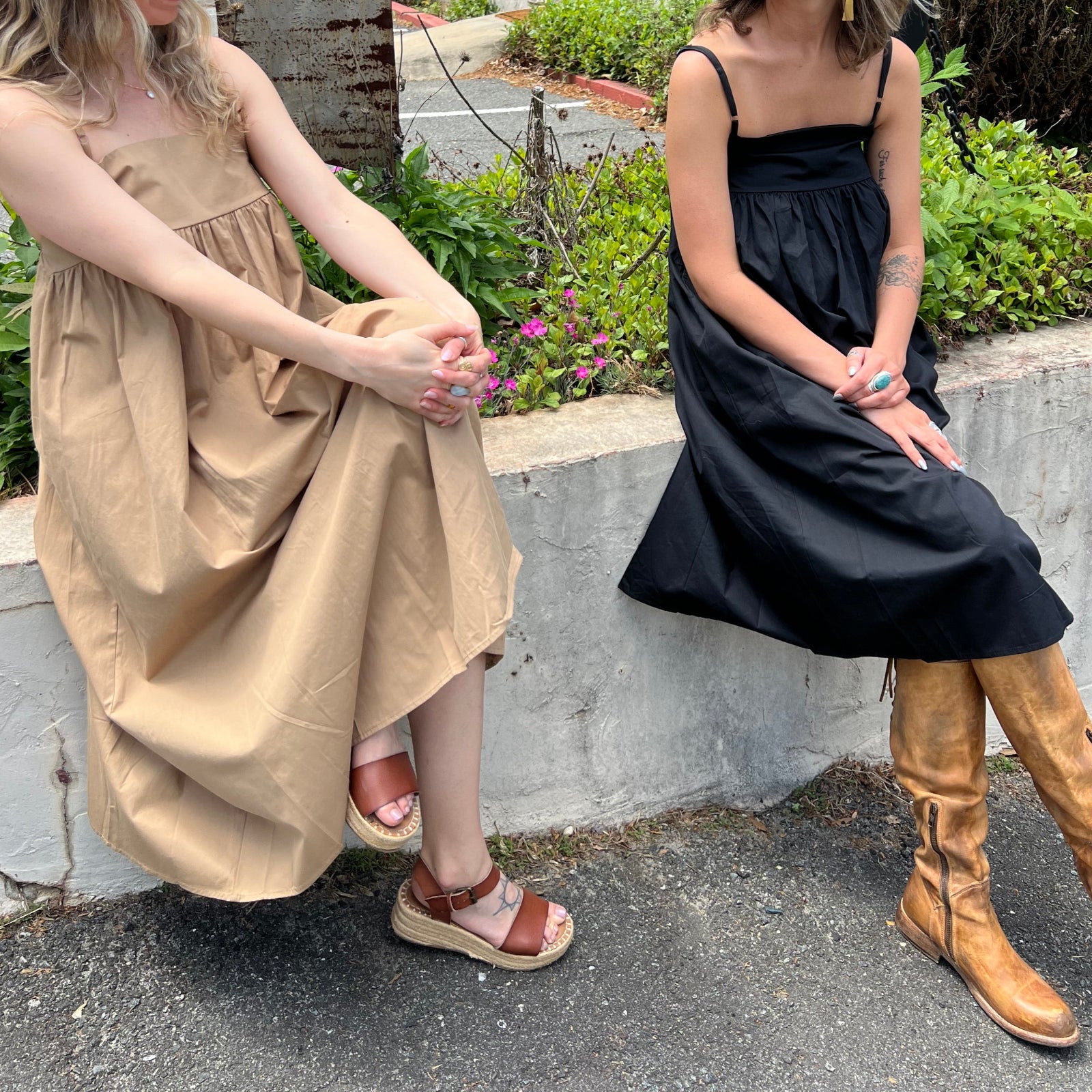 Two women wearing the Solid Color Pleats Suspender Acetate Fiber Free Size Dresses in black and khaki while sitting on a white ledge in front of greenery.