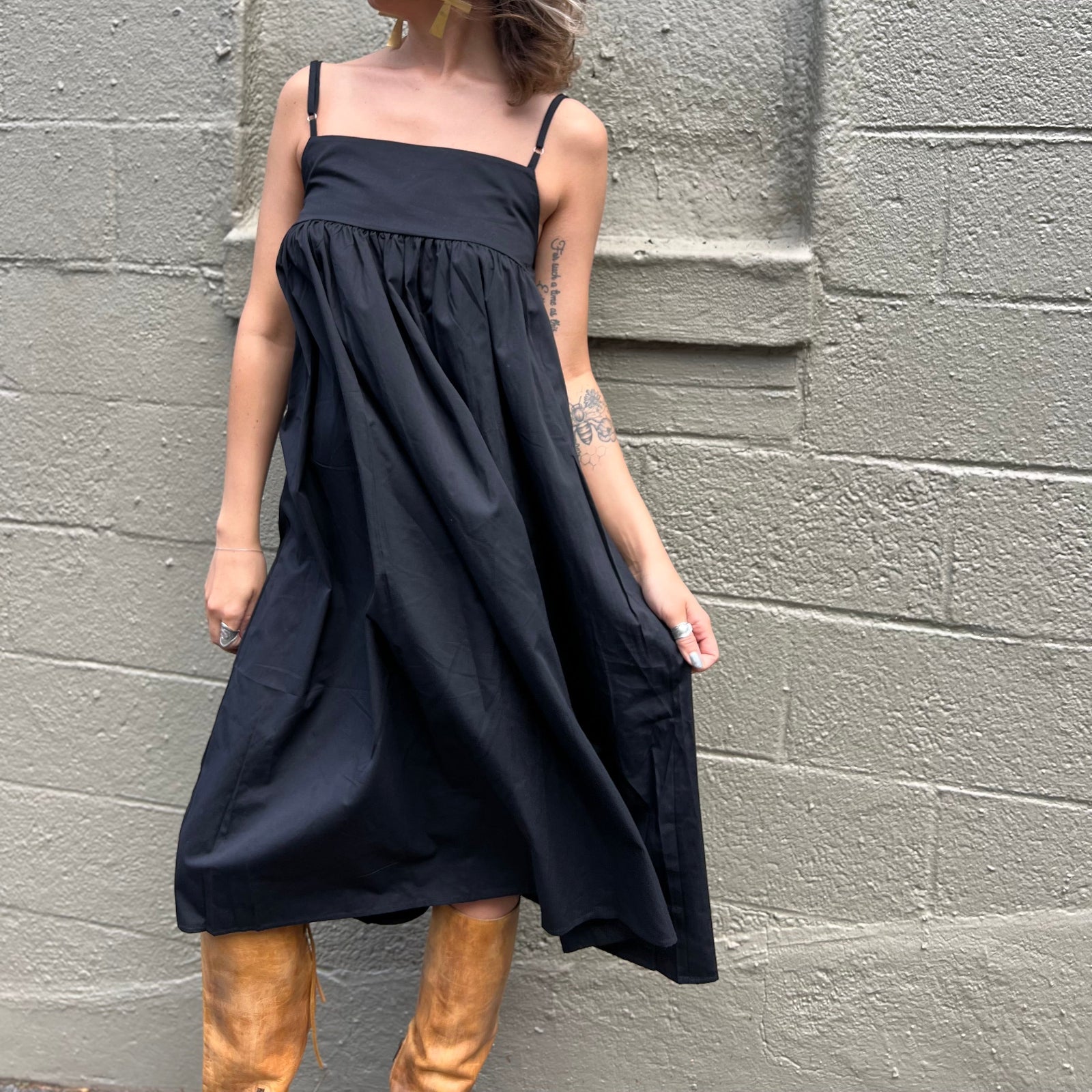 A woman wearing the black Solid Color Pleats Suspender Acetate Fiber Free Size Dress in front of a white brick wall.
