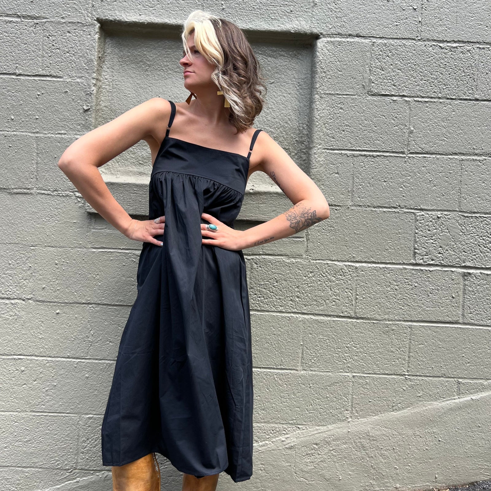 A woman wearing the black Solid Color Pleats Suspender Acetate Fiber Free Size Dress in front of a white brick wall.