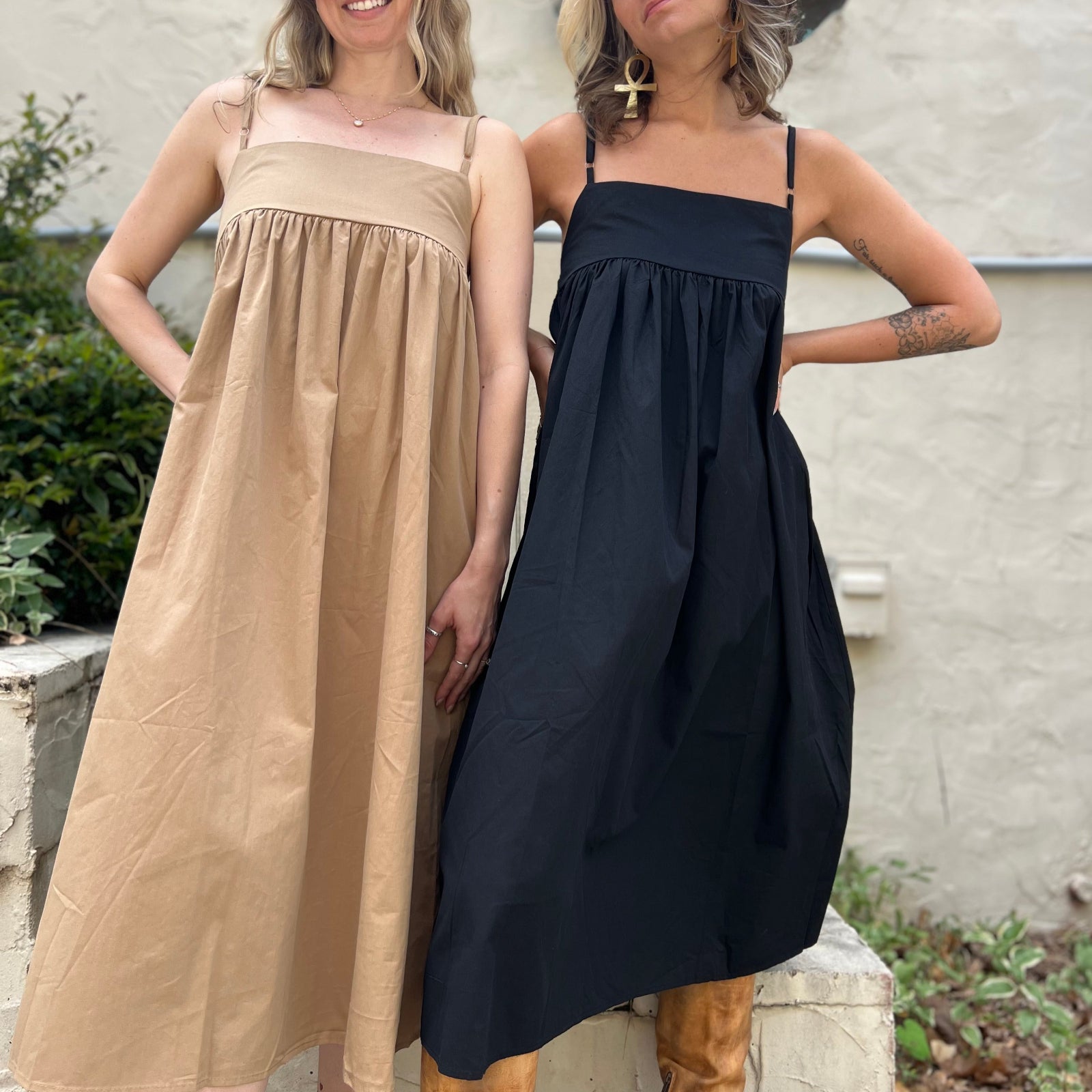 Two women wearing the Solid Color Pleats Suspender Acetate Fiber Free Size Dresses in black and khaki in front of a white wall with greenery.