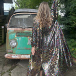 A woman wearing the Rainbow Rage Sequin Cocoon Jacket with a black dress in front of a vintage teal Volkswagen bus, showing off the back.