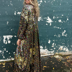 A woman wearing the Rainbow Rage Sequin Cocoon Jacket with a black dress in front of a blue distressed wall, showing off the side.