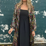 A woman wearing the Rainbow Rage Sequin Cocoon Jacket with a black dress in front of a blue distressed wall.