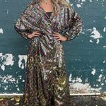 A woman wearing the Rainbow Rage Sequin Cocoon Jacket with a black dress in front of a blue distressed wall.