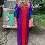 A woman wearing the Vintage Rainbow Sequin Striped Long Kimono with a black dress in front of a vintage teal Volkswagen bus, showing off the back.