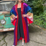 A woman wearing the Vintage Rainbow Sequin Striped Long Kimono with a black dress in front of a vintage teal Volkswagen bus.