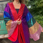 An up-close image of a woman wearing the Vintage Rainbow Sequin Striped Long Kimono with a black dress in front of a vintage teal Volkswagen bus.