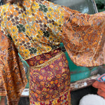 A woman the Hippie Golden Patchwork Kimono Robe Duster with a white bralette and red velvet bellbottoms in front of a vintage Volkswagen bus.