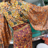 A woman the Hippie Golden Patchwork Kimono Robe Duster with a white bralette and red velvet bellbottoms in front of a vintage Volkswagen bus.