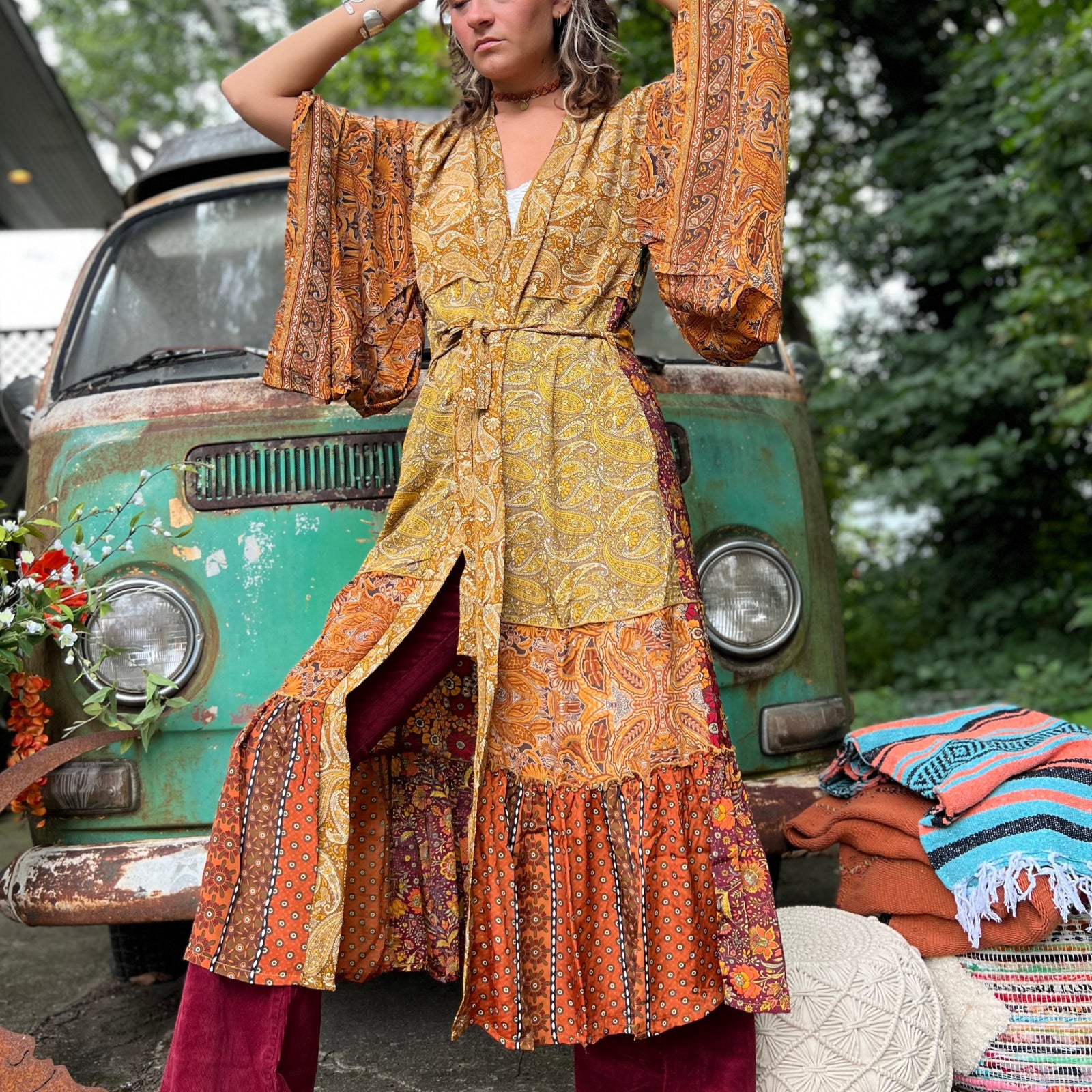 A woman the Hippie Golden Patchwork Kimono Robe Duster with a white bralette and red velvet bellbottoms in front of a vintage Volkswagen bus.
