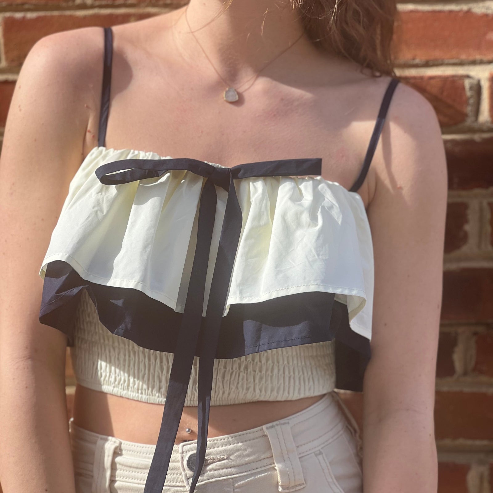 A woman wearing the Contrast Cotton Flare Bow Top with white jeans in front of a brick wall.