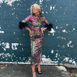 A woman wearing the Catroux dress in Hysteria with a pair of heels in front of a dark teal wall.