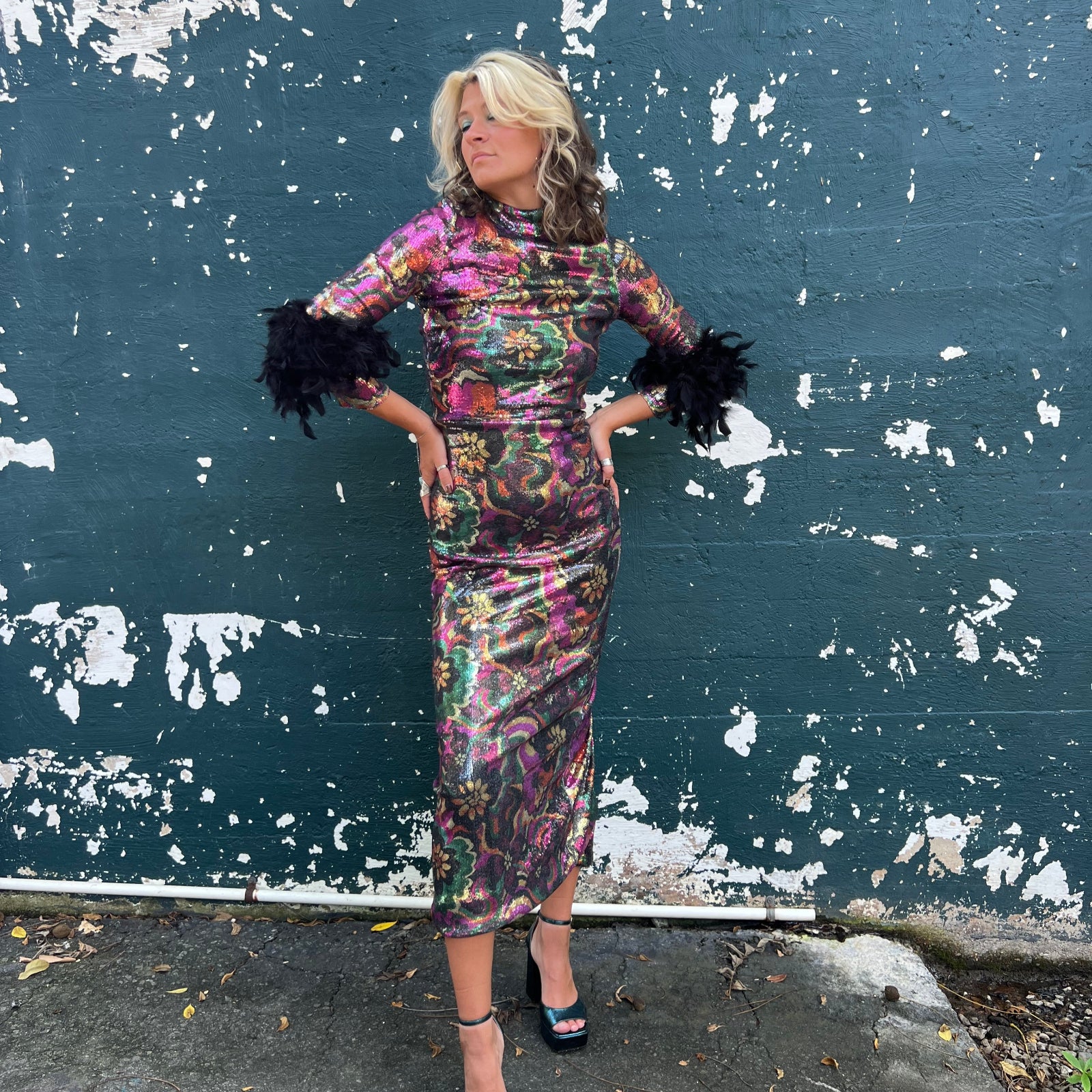 A woman wearing the Catroux dress in Hysteria with a pair of heels in front of a dark teal wall.
