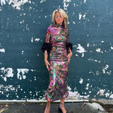 A woman wearing the Catroux dress in Hysteria with a pair of heels in front of a dark teal wall.