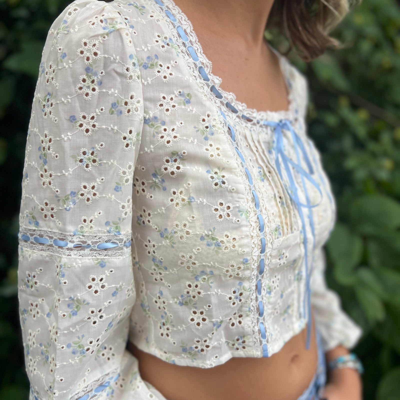 A woman wearing the blue Eyelet Floral Ribbon Trim Cotton Crop Top with jean shorts in front of some greenery.