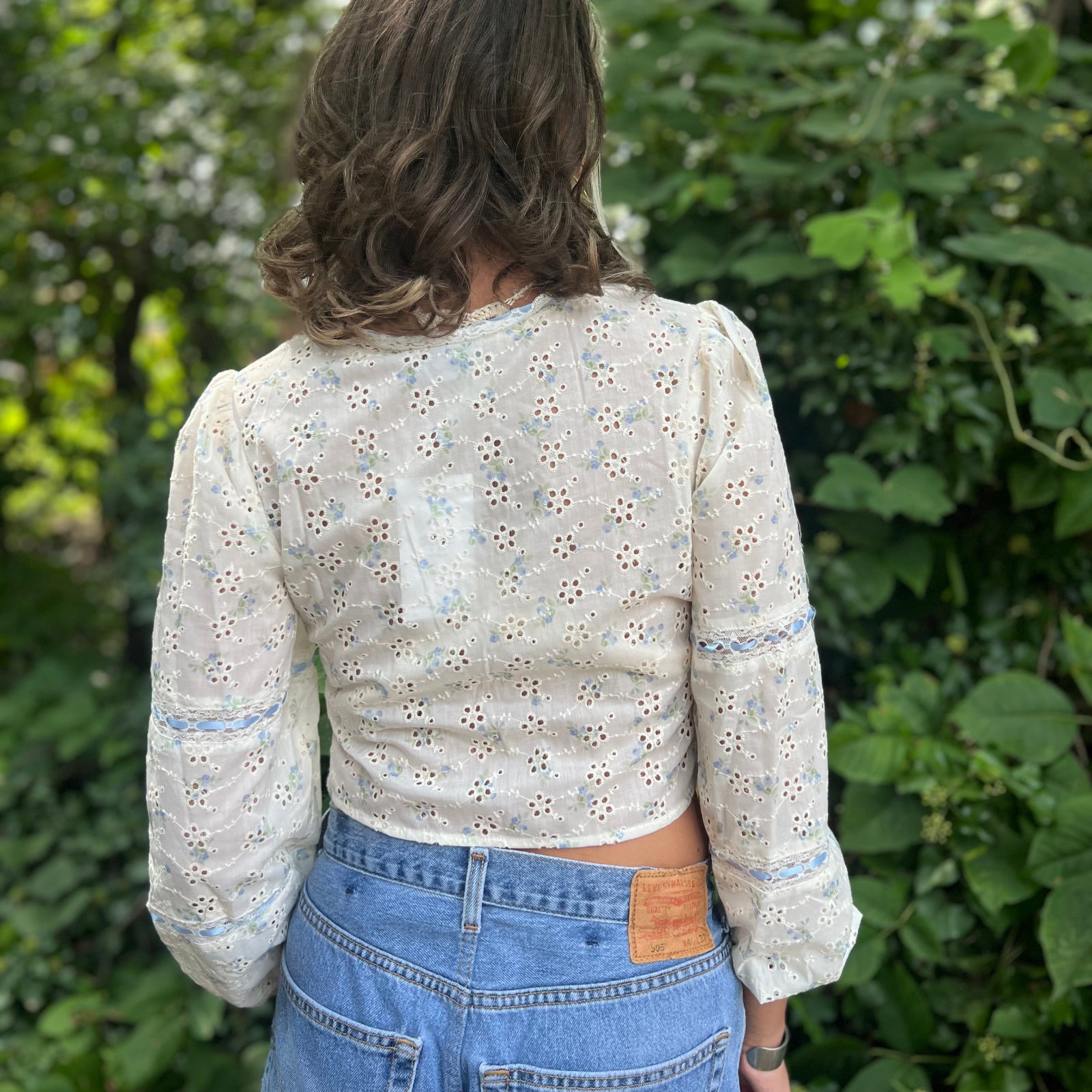 A woman wearing the blue Eyelet Floral Ribbon Trim Cotton Crop Top with jean shorts in front of some greenery.
