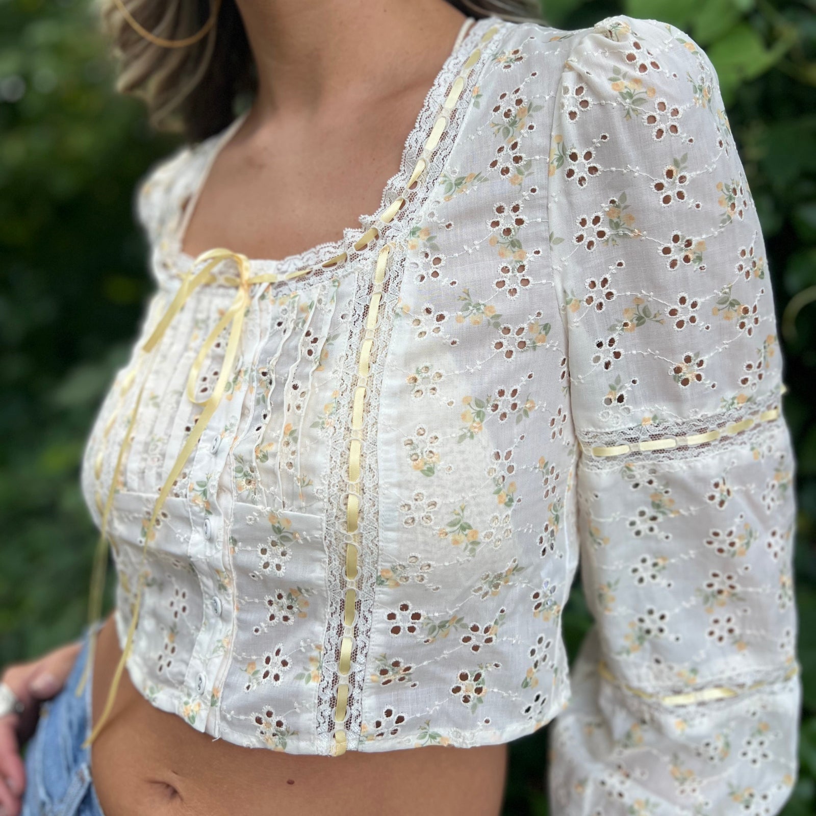 A woman wearing the yellow Eyelet Floral Ribbon Trim Cotton Crop Top with jean shorts in front of some greenery.