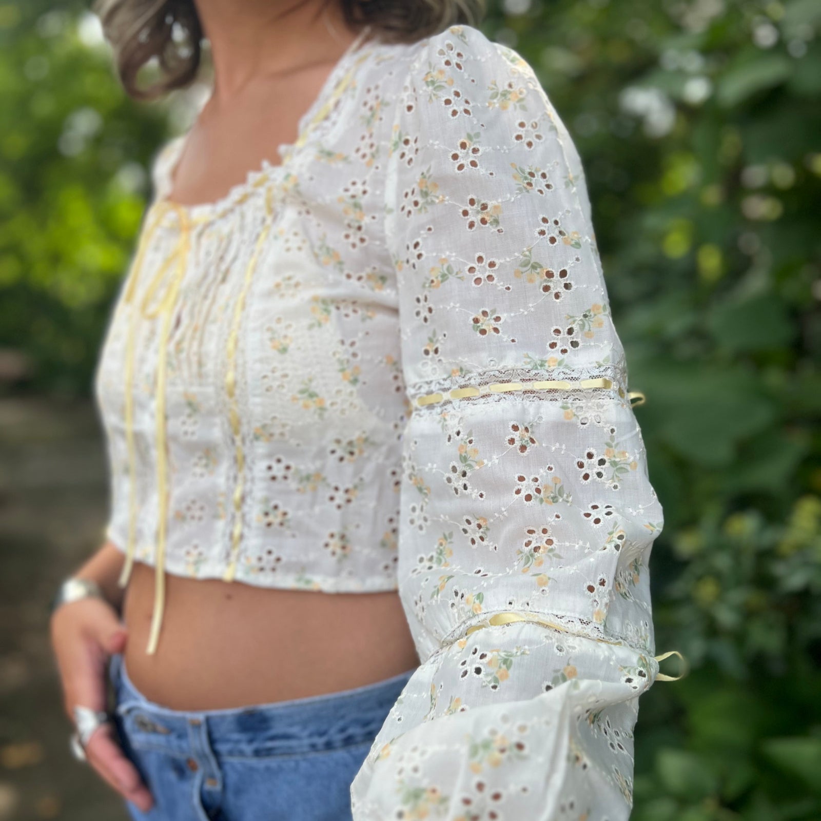 A woman wearing the yellow Eyelet Floral Ribbon Trim Cotton Crop Top with jean shorts in front of some greenery.