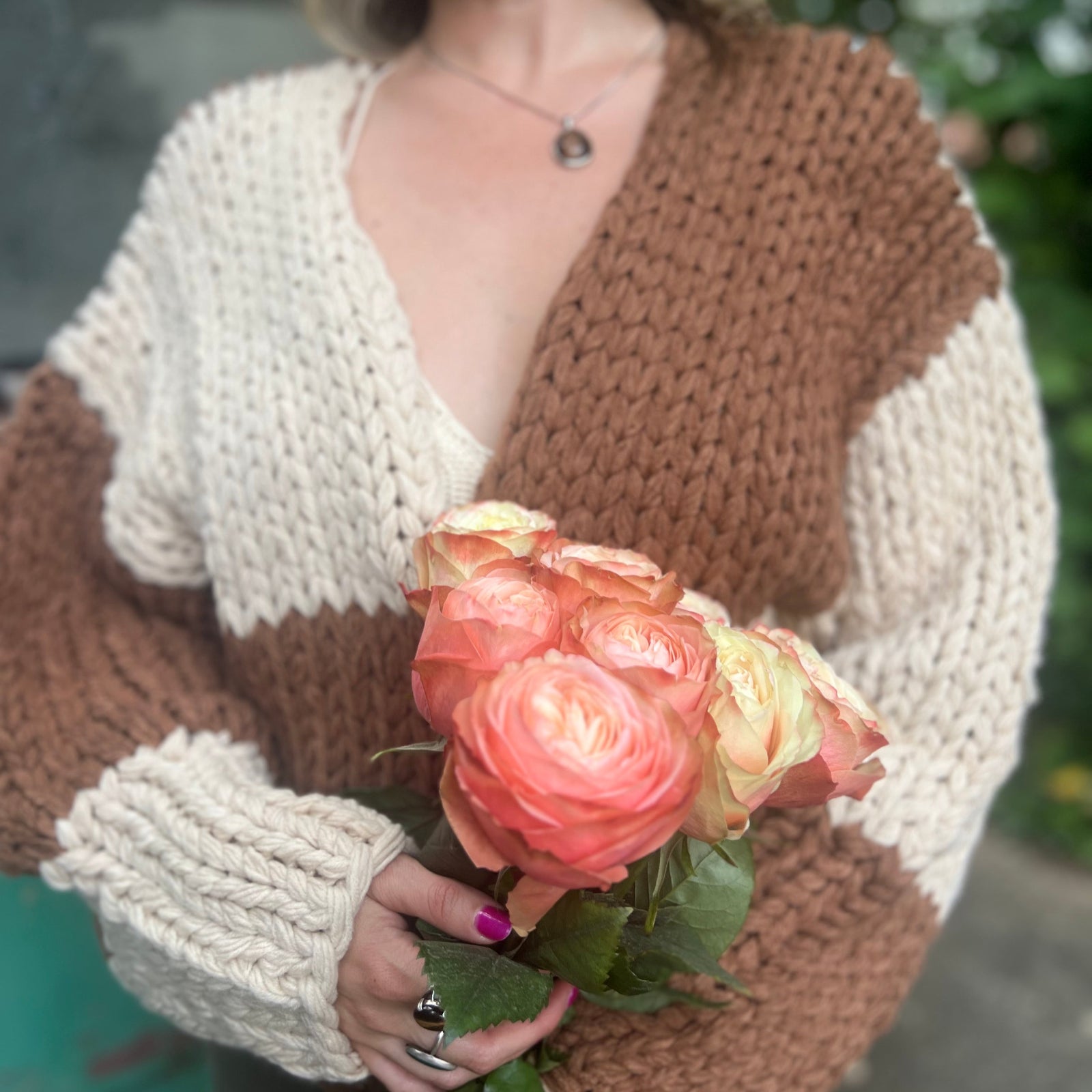 A woman wearing the Cream and Brown Color Block Chunky Knit Open Front Sweater Cardigan with a white crochet top while holding flowers.