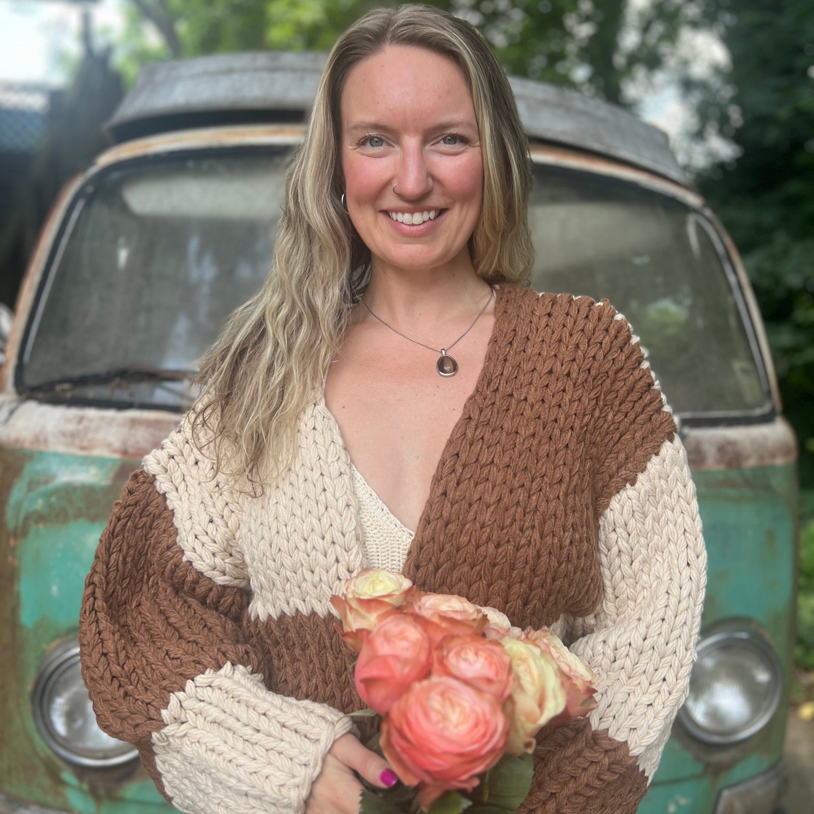 A woman wearing the Cream and Brown Color Block Chunky Knit Open Front Sweater Cardigan with a white crochet top while holding flowers.