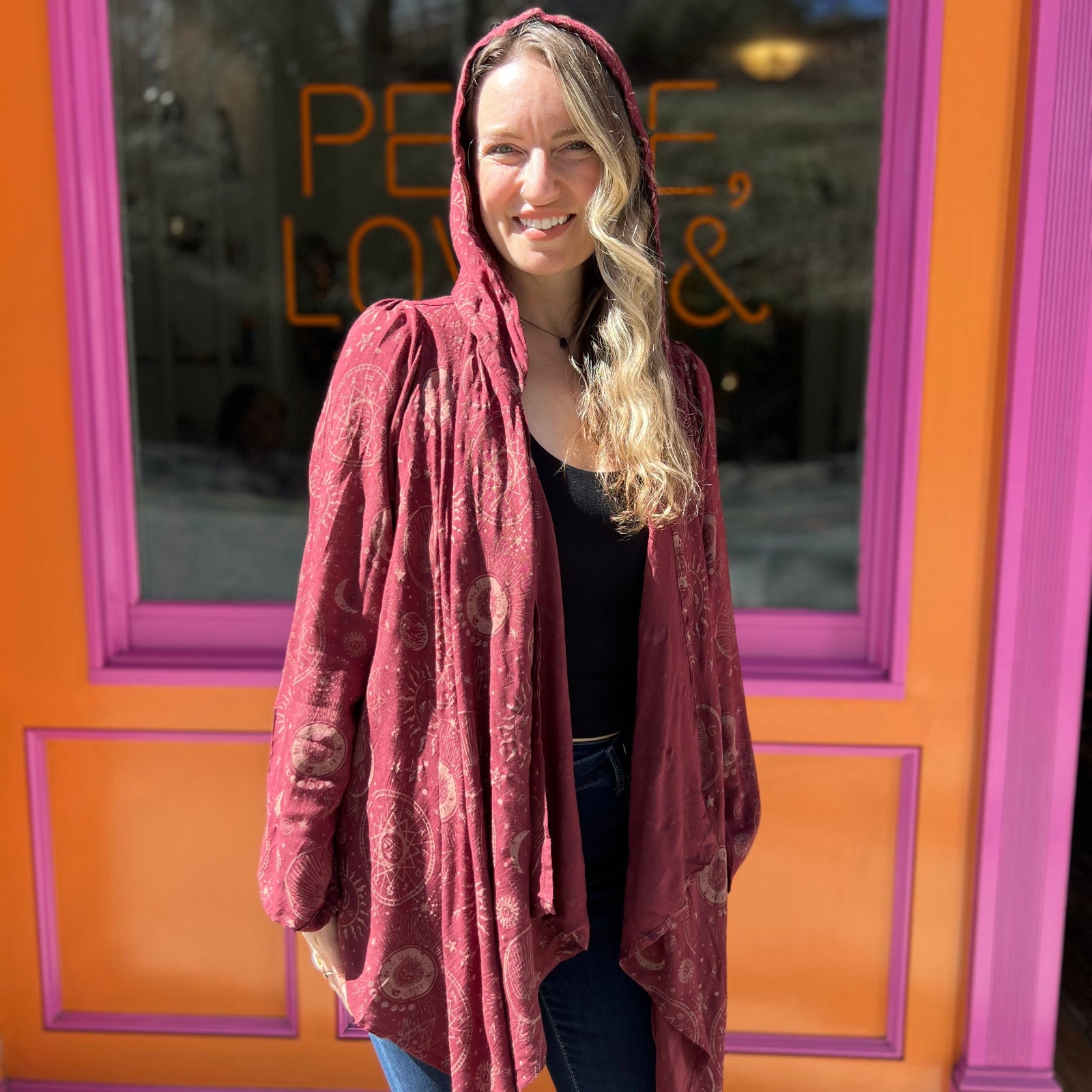 A woman wearing the Flowy Hoodie Waterfall Hooded Cardigan with a black tank and bell bottoms in front of a pink and orange wall.