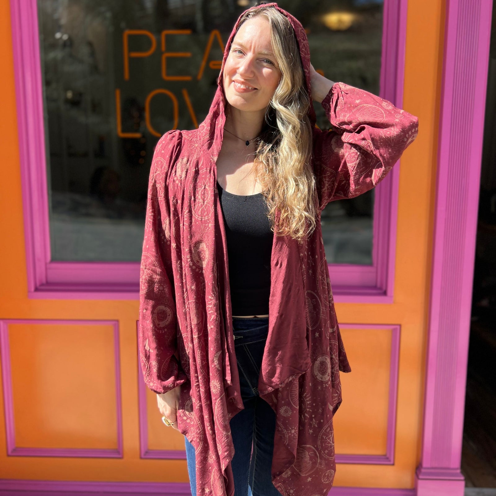 A woman wearing the Flowy Hoodie Waterfall Hooded Cardigan with a black tank and bell bottoms in front of a pink and orange wall.
